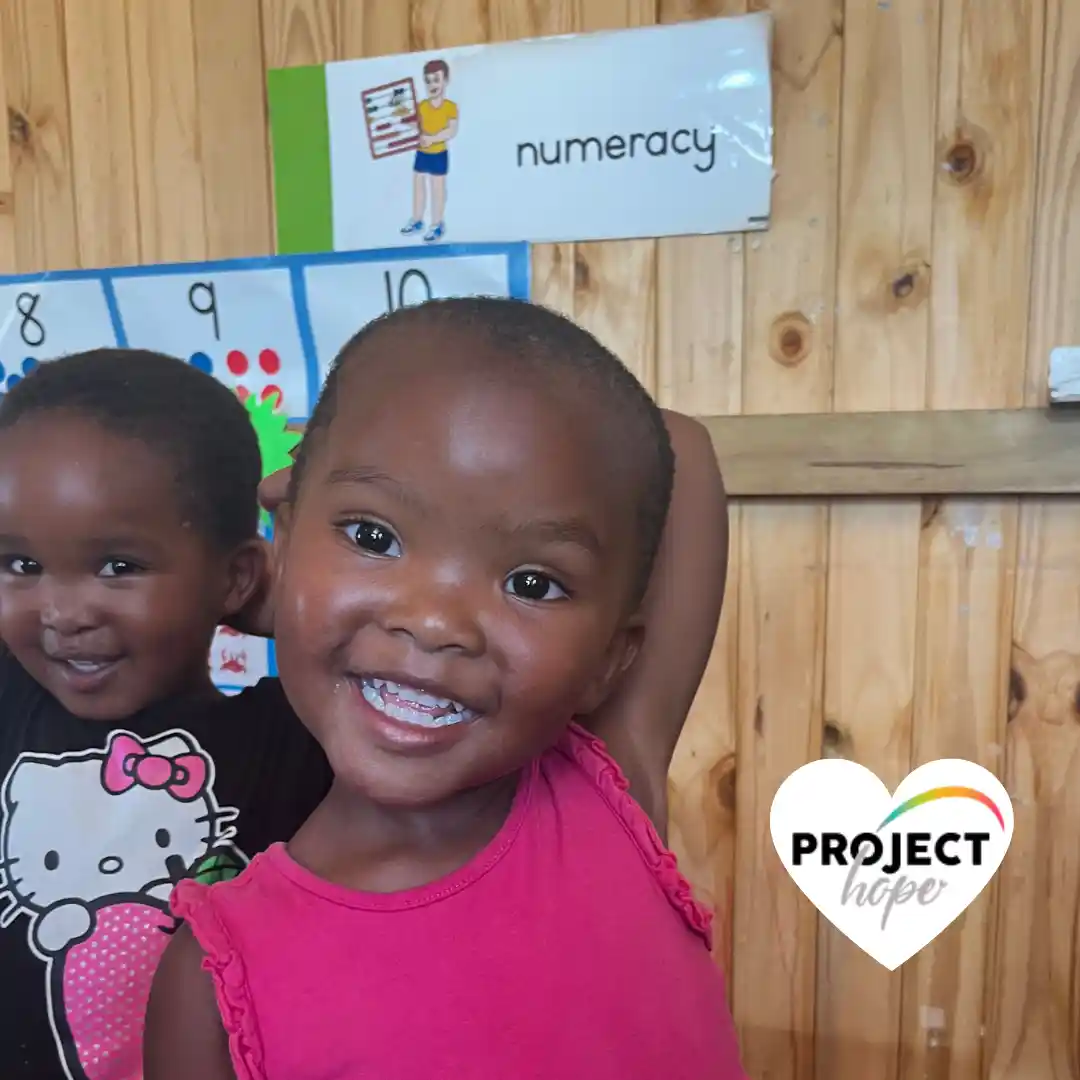 Two toddlers smile in a classroom in front of a "numeracy" sign and educational posters. 8. A group of under privileged children gathered together outdoors on play mats at a Project Hope funded daycare in South Africa.