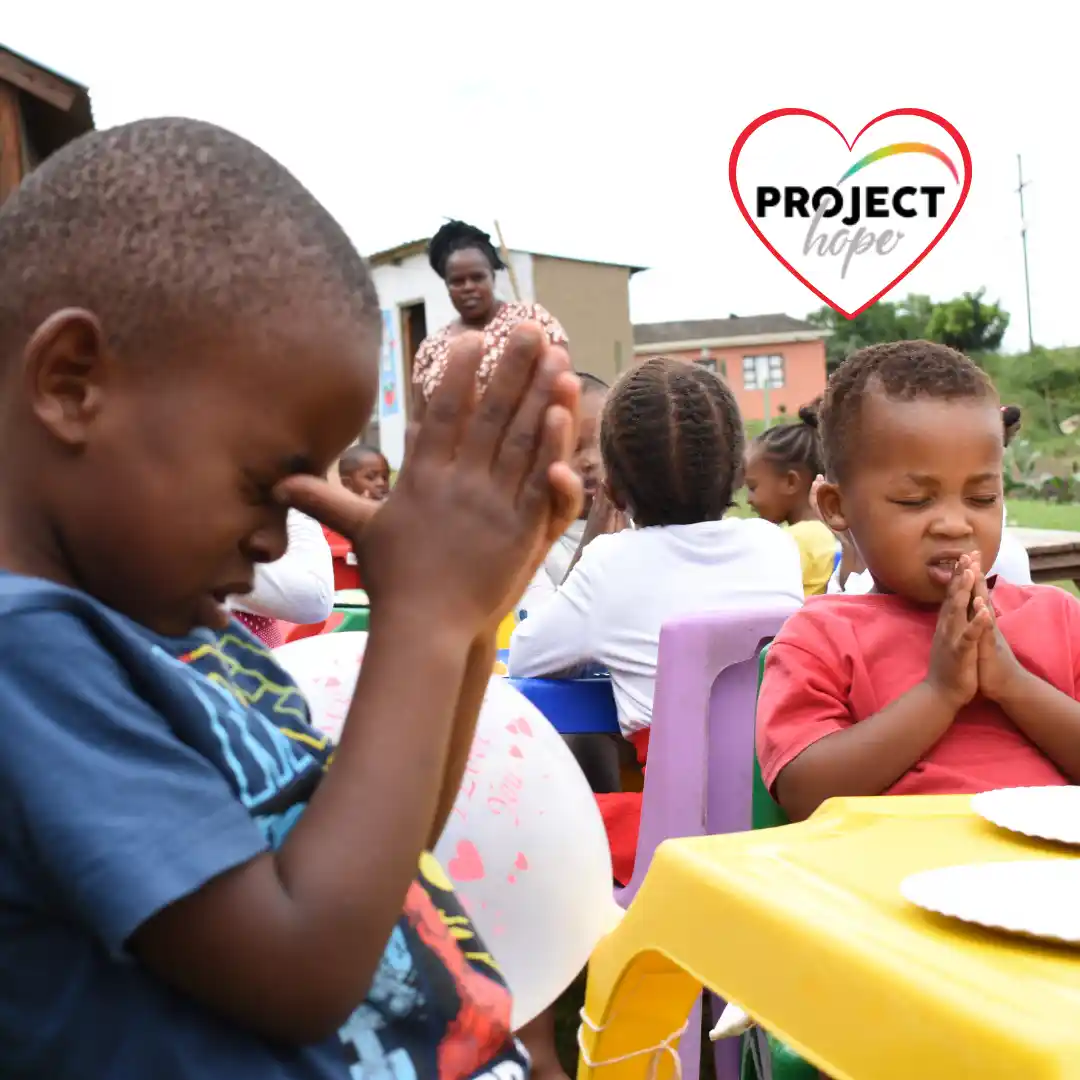 meals-donate-to-early-childhood-development Young preschool students bow their heads and clasp their hands in prayer before eating nutritious school meals at outdoor tables.
