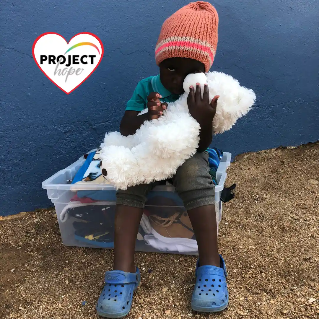 A young Sanele sits on a plastic storage container hugging a white teddy bear on the day he received sponsorship from Project Hope’s ECD programme.