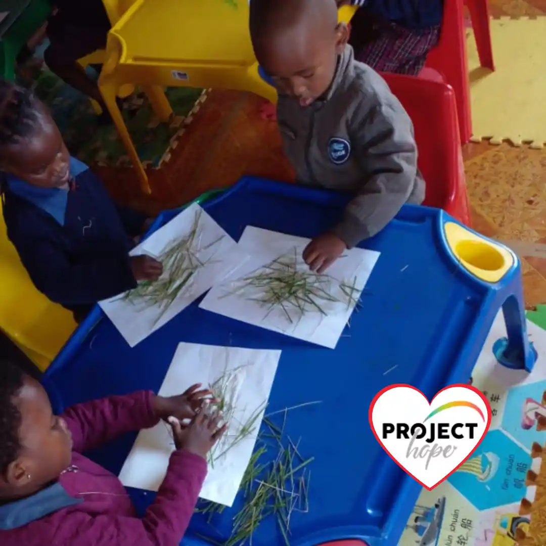 Young children arrange grass on sheets of paper at a classroom table during an interactive lesson using a play-based curriculum.